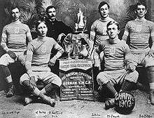A black-and-white image of young male basketball players dressed in team uniform, sitting around a display that holds trophies. The plaque below the display reads "Pan-American Basket Ball Champions."