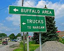 Two green signs on metal poles with white lettering and arrows pointing to the left. The larger one says "Buffalo area" and the smaller says "Trucks to Warsaw". There is a road behind it to the left leading to an intersection at the rear.