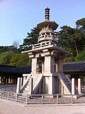 A stone pagoda with elaborated tiers, a small lion status, and stairs. Blue skies and a roof of a building and trees are shown on the background