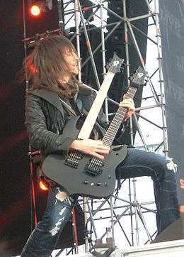 A long brown haired man in a black leather jacket and torn up blue jeans plays a black double necked guitar on a stage.