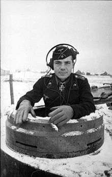 A black-and-white photo of a man protruding from the cupola of a tank wearing a black military uniform, side cap and headset.