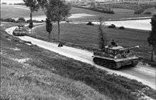 Four tanks move down a tree lined lane in open country.