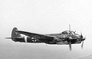 A twin engine propeller powered aircraft in flight and viewed from the right side. The aircraft bears multiple markings including a black and white cross on its side and swastika on the tail fin.