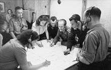 Black-and-white photograph of men and women wearing headsets standing around a table and making notes on a large map.