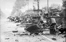 Several wrecked vehicles along the verge of a tree and hedge lined road. A destroyed gun, twisted metal and debris occupy the foreground.