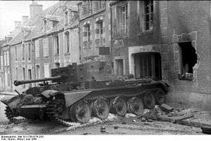 A tank, with debris strewn around it, is in front of a damaged and fire scorched house. The tank is position on the pavement and partially on the road.