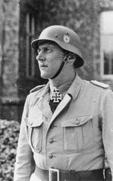 A man wearing a military uniform and steel helmet with an Iron Cross displayed at the front of his uniform collar.