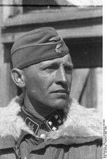 A black-and-white photograph of a man wearing a military uniform with fur collar, side cap and a neck order in shape of an Iron Cross. His cap has an emblem in shape of a human skull and crossed bones.