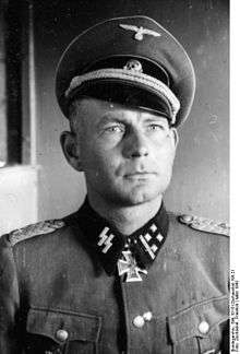 Black-and-white photo of a man wearing a peaked cap, military uniform with an Iron Cross displayed at his neck.