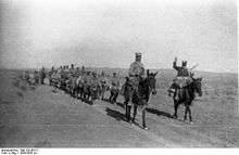 A column of around a dozen Foreign Legion troops on foot, followed by a similar number mounted on donkeys and led by two mounted officers/NCOs proceeding along a road