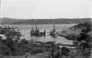 A large warship is tied to the dock in a narrow channel of water. Two smaller ships are alongside her.