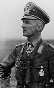 Black-and-white portrait of a man in semi profile wearing a peaked cap, military uniform with an Iron Cross and binoculars suspended from his neck.