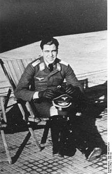 A man sitting in folding chair wearing a military uniform, a peaked cap is resting on his knees.