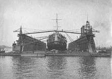 Large ship atop a floating platform with two tall walls running along her side, and construction equipment between the walls and the ship