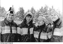 Three men and a woman are lined up side-by-side wearing winter caps and identical winter jackets. They are outdoors and snow is falling.