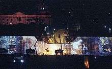 A wall covered in graffiti with a circular movie screen and lights atop it. Several cranes are visible in the center.