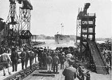 A large ship slides into the water. A tall crane stands on one side and a large scaffolding on the other. A crowd of spectators has gathered all around.