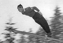 A man, in ski suit, is in flight of a ski jump, he is leaning forwards, in the middle of his jump.