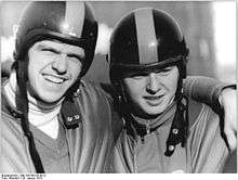 Two men wearing helmets embrace each other. The man on the left has his left arm over his partner's shoulder and grins while protecting his eyes from the direct sunlight with his brows.