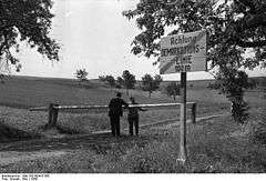 Two people stand either side of a lowered border pole on a dirt road with a sign in the foreground