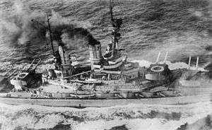 Overhead view of a large battleship; black smoke pours from its smoke stacks as it steams through choppy seas.