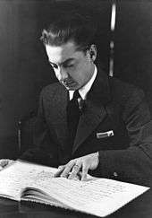 young man with slicked back dark hair, looking down at a musical score