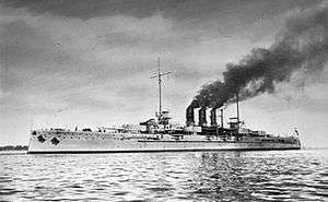 Large gray battleship at sea. Dark smoke streams back from its three closely arranged funnels.
