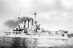 A large light gray battleship sits in harbor, smoke drifts up from its three tall funnels