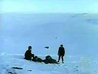 Four men in a bleak icy landscape. The man on the left is sitting, the two middle men are lying down and the man on the right is standing.