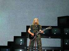 Madonna wearing olive green clothes sings to a microphone while holding a black electric guitar in her hands.