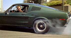 Photograph of a car with a driver looking backwards out of its window. The car's rear tire is smoking because it is spinning against the road.