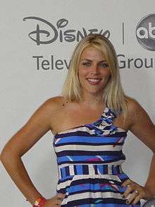 A woman with shoulder-length blonde hair and wearing a patterned dress poses during an event.