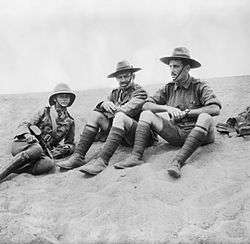 An informal black and white group portrait of three men in military uniform. They are sitting on the ground in what appears to be a desert.