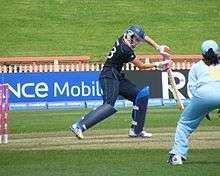 View of cricket pitch; side profile of a woman in dark blue who is batting and back of another woman wearing light blue and fielding
