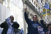 A black man on the left wearing a white T-shirt and dark jacket smiles holding a sign, while a white man on the right wearing a blue sweatshirt which reads "Champions" points upwards. White streamers hang from a building in the background.
