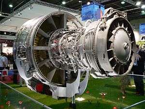 An exposed jet engine at a trade show. The rear of the polished metal fan case is visible on the left. The outer casing of the compressor section, covered in fuel lines and electrical wires is to the right of the fan case. The right of the image shows the back of the engine, the exhaust area of the turbine section.