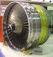 The front fan of a jet engine facing the left of the image, surrounded by its metal casing. The conical inlet in seen right in front of the metal fan blades. The fan casing is seen in three distinct (but attached) sections from left to right, first a silver-colored section, then a golden-colored section, then another silver-colored section.