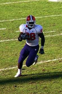 Football player in #28 white uniform with blue pants and red helmet runs with the football