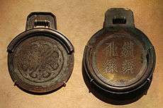 Two circular bronze plates with square handles. The plate on the right is thicker, has four characters embossed into it, and contains a rim around about three fourths of the edge of the plate. The plate on the left is thinner, and contains a rim that, when the two plates were stacked on one another, would perfectly meet the edges of the first plate.