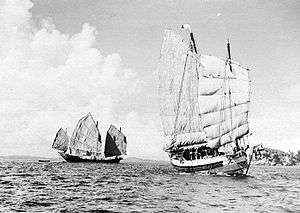 Black and white view of the ocean with an island visible on the horizon to the right. A sailing ship on the left (three sails visible) shows the full length of its hull while another on the right (two sails visible) shows its forward bow.