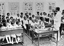 An adult man speaks to several dozen children who are seated on school benches. Behind them on the wall are hanging posters containing various diagrams.
