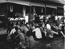 Several dozen men are in squatting positions in front of building. Inside, men dressed in white are sitting behind tables and standing by.