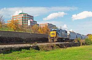 A blue diesel locomotive, with "CSX" in blue letters on its yellow front, pulling several hopper cars along a railroad track. In the background are some postmodernist brick office buildings with green spired roofs.