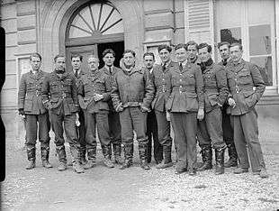 Thirteen men in dark military uniforms standing outside a building
