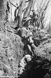 A soldier wearing a slouch hat stands in a trench on the side of a hill, while other soldiers move in the background amidst the trees. In the bottom of the trench is the body of another soldier