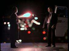 Two men both dressed in black suits standing in the middle of a road at night. The man on the right, is holding a gun and is aiming on the man on the left. Behind them, bright city lights and a car.