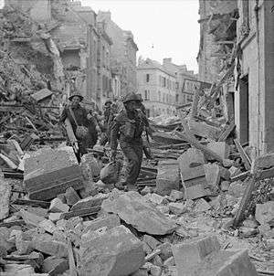 A line of soldiers carefully clambering over the heaped rubble of destroyed buildings in a badly-damaged street