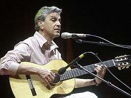A white man with gray hair, wearing a striped shirt with white and pink colors, holding an acoustic guitar with two microphones in front of him.