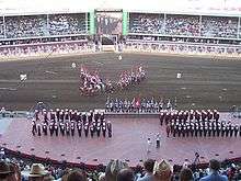 Approximately 50 people in red, black and white uniforms stand on a stage as a team of riders on horseback carry Canadian Flags in the background.