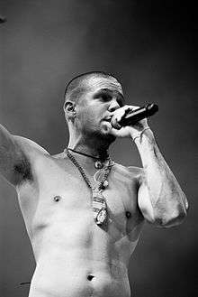 A black and white image with a white male, shaved, shirtless, holding a microphone with his right hand while lifting his left hand. On his neck hangs a necklace.